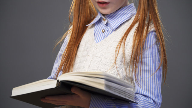Close up redhead girl in a blue shirt and a white vest  with two pigtails holding a book in the hands, gray background