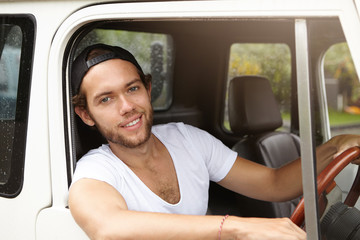 Young Caucasian traveler wearing casual t-shirt and baseball cap driving his white sports utility vehicle, enjoying road trip and summer vacations, sticking his head out of open window and smiling