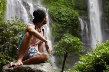 Nature, wildlife and travel concept. Young barefooted hiker wearing snapback sitting on big stone and enjoying beautiful view around him. Hipster relaxing deep in rainforest, looking at waterfall