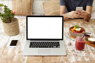 Front view of workplace of self-employed woman or freelancer: generic laptop computer resting on wooden table with smart phone, food and glass of smoothie. Man in background using electronic device