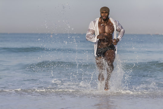 Dark Muscular Man With Wet White Shirt Running And Splashing Water Out Of Ocean 