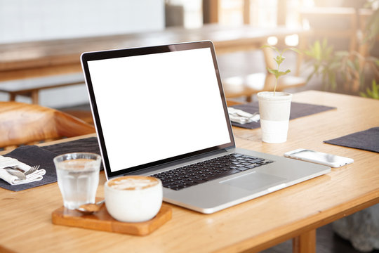 Modern technology and communication concept. Close up view of open notebook computer, generic smart phone and mug of cappuccino resting on wooden table, against blurred interior of cafeteria