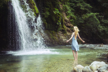A woman standing in the river with her  Raised hands to waterfall.