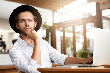 Indoor shot of handsome young blogger in headwear working on new post for his blog using wi-fi on generic laptop pc, holding hand on his chin and looking ahead of him with thoughtful expression