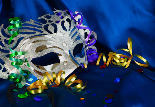 Shiny silver Mardi Gras mask with gold, green and purple ribbons and confetti all on a luxurious blue satin draped background. Copy space.