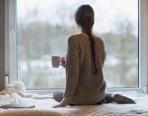 Thoughtful young brunette woman with book and cup of coffee looking through the window, blurry winter forrest landscape outside - 132533519