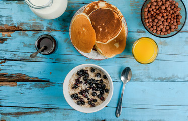 Blue rustic table with everything you need for a family continental breakfast: milk, chocolate cereal balls, oatmeal with berries, pancakes, jam and orange juice, top view