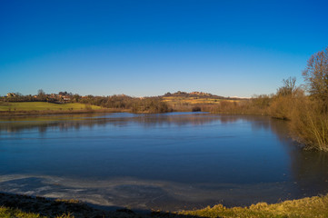 expanse of green grass on the lake with blue sky, countryside and farm around the lake, sunlit water