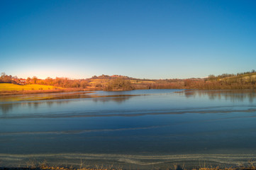 expanse of green grass on the lake with blue sky, countryside and farm around the lake, sunlit water