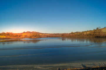 expanse of green grass on the lake with blue sky, countryside and farm around the lake, sunlit water