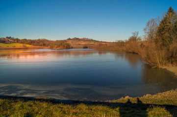 Naklejka premium expanse of green grass on the lake with blue sky, countryside and farm around the lake, sunlit water