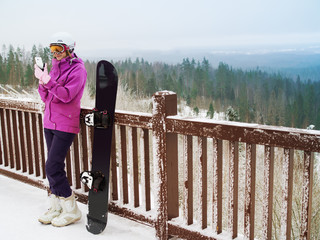 Attractive girl on mountain with snowboard. Snowboarder with mobile phone in hand. Girl snowboard helmet phone suit sunglasses.