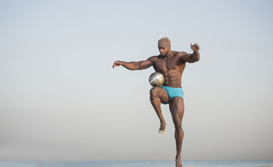 Fototapeta na wymiar muscular man in blue shorts playing with soccer ball on beach