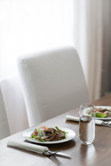 An oil table set for lunch. A white plate with buckwheat, mushrooms, arugula and prosciutto. A glass of water. White upholstered chairs and white curtains in the background