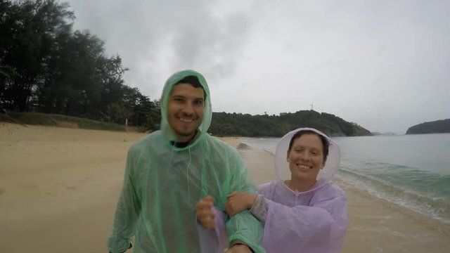 Loving Couple Walking and Smiling on the beach in raincoats. Rain season Thailand. Selfie Action on camera.