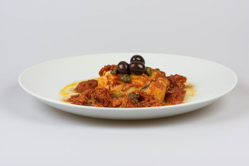 Italian codfish with black olives, cappers and tomato in a design plate, isolated on a white background