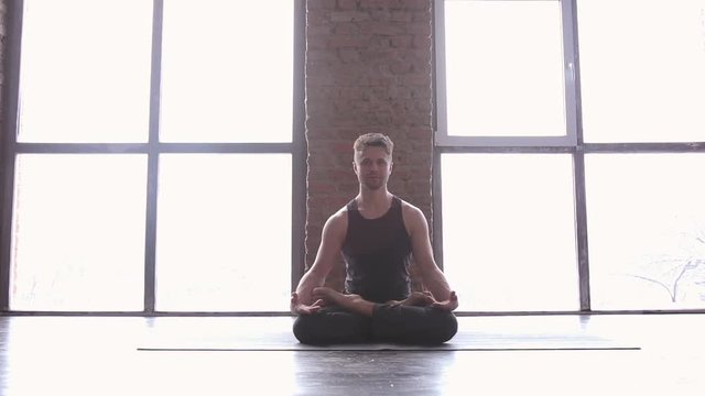 Portrait of a handsome man practicing meditation and yoga against an urban background with picture window and red brick wall on black wooden floor. Yogi men sitting in lotus pose and meditates
