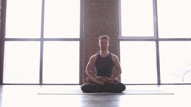 Portrait of a handsome man practicing meditation and yoga against an urban background with picture window and red brick wall on black wooden floor. Yogi men sitting in lotus pose and meditates