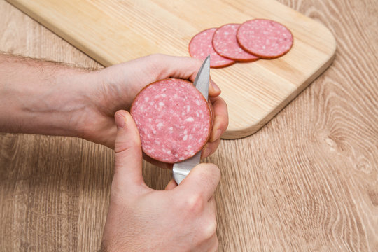 Man's hands on the wooden board with a knife cutting a smoke-dried sausage in the kitchen.