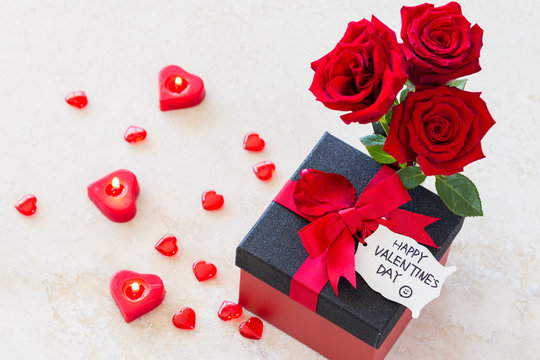 Valentine gift/Valentines concept with bouquet of roses, heart shaped candles and wrapped gift with “happy valentine’s day” note on wooden table