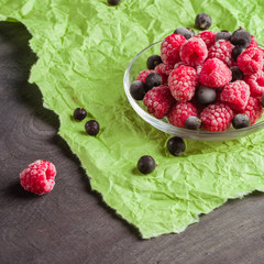 Frozen raspberries in a glass saucer. Frost on the berries. Dark and green background. Green crumpled paper.