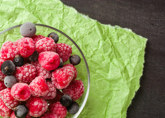 Frozen raspberries in a glass saucer. Frost on the berries. Dark and green background. Green crumpled paper.