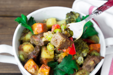 closeup beef meat stewed with vegetables in ceramic pot with a piece on a fork on wooden background