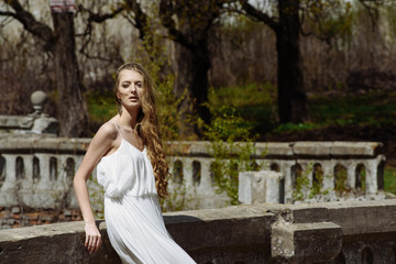 Outdoor summer portrait of young pretty cute girl. Beautiful woman posing at old bridge.  in white dess standing near stone railing. Photo has an  model.