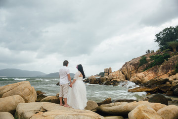 groom in white suit and the bride in a dress holding each other's hands  standing on the background of the sea. beautiful landscape with big stones. concept wedding day