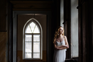 Fototapeta premium Indoor summer portrait of young pretty cute girl. Beautiful woman posing beside fairytale window inside wood cabinet, scars old castle. Bride in white atlas dress. Photo has a model.