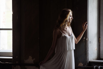 Obraz premium Indoor summer portrait of young pretty cute girl. Beautiful woman posing beside two fairytale windows inside wood cabinet, scars old castle. Bride in white atlas dress. Photo has a model.
