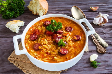 Tomato soup with orzo and smoked sausages in white casserole on wooden rustic table. Fresh bread...