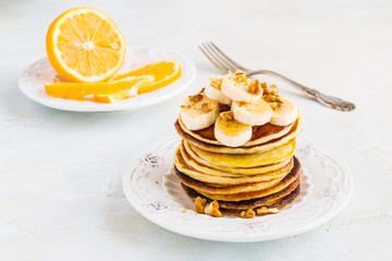 Stack of homemade pancakes with banana, maple syrup and walnuts on vintage plate. Fork, fresh sliced lemon, white and gray concrete background.