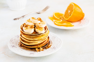 Stack of homemade pancakes with banana, maple syrup and walnuts on vintage plate. Fork, fresh sliced lemon, white and gray concrete background.