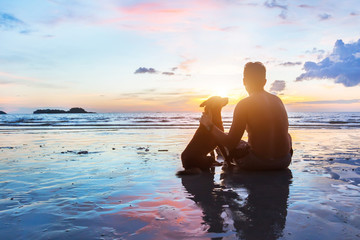 friendship concept, man and dog sitting together on the beach at sunset - Powered by Adobe