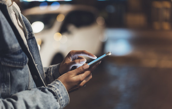 Girl pointing finger on screen smartphone on background auto taxi, illumination bokeh color light in night atmospheric city, hipster using in hands and texting mobile phone, mockup street lifestyle