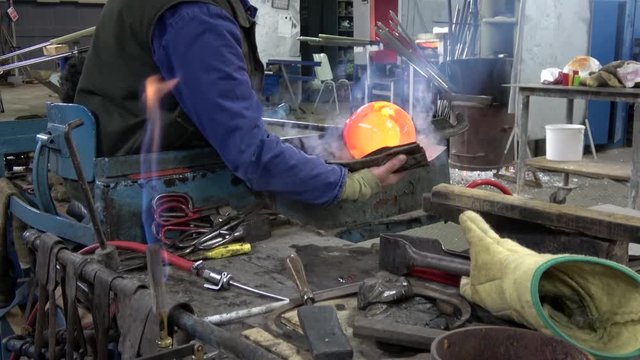 Glassblowing workshop showing glassblower gaffer spinning rod with glowing hot liquid glass on work bench using wet sandpaper to shape liquid glass crystal amazing colors warm yellow orange 4k quality