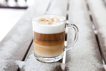 Cup with cappuccino coffee on the wooden bench covered with snow. Blurred winter background with snowflakes.
