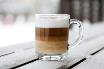 Cup with cappuccino coffee on the wooden bench covered with snow. Blurred winter background with snowflakes.
