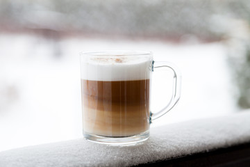 Cup with cappuccino coffee on the wooden bench covered with snow. Blurred winter background with snowflakes.
