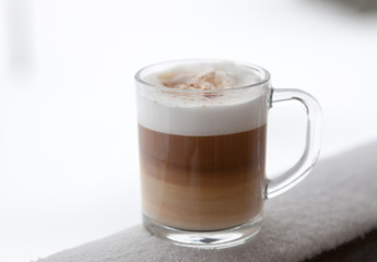 Cup with cappuccino coffee on the wooden bench covered with snow. Blurred winter background with snowflakes.
