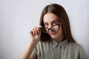 A portrait of pretty young girl wearing big glasses and shirt having cunning expression looking from under glasses holding her hand on glasses. An isolated attractive girl with furtive look