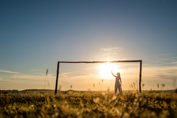 Fototapeta na wymiar Young romantic woman on the meadow at sunset. The girl in the white dress enjoys the outdoors. Warm light . Strong glow of the sun behind the girl . The girl inside the frame vintage football gate
