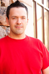 Confident man looks at the camera.  Caucasian man wearing a red shirt with dark hair leaning against a stone wall outside.