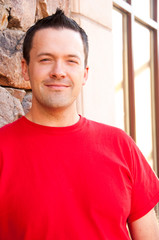 Obraz premium Young man leans against a building with a rock wall and smiles at the camera. Man wears a red tee shirt, dark hair, caucasian man in this late 20s.