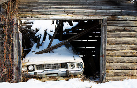 An abandoned vintage car backed into a log shed with a collapsed roof onto the car in winter