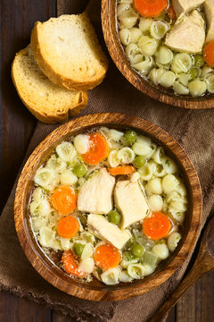 Homemade chicken soup with pea, carrot and small shell pasta in wooden bowls, toasted bread on the side, photographed overhead with natural light (Selective Focus, Focus on the top of the soups)