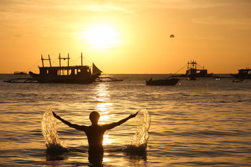 Silhouette of man on the beach splashing water with arms wide open and sailboats on the background at sunset in Boracay White Beach, Philippines
