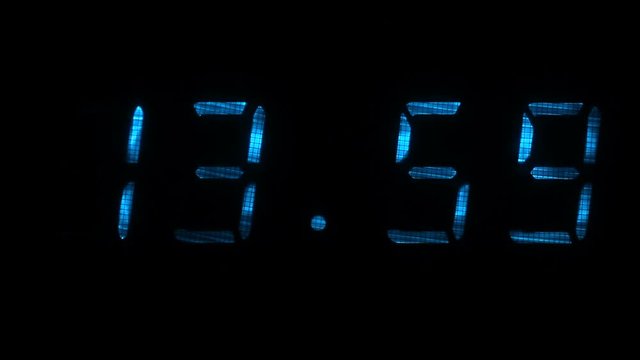 Digital clock with fluorescent display shows the time of 13 hours 59 minutes to 14 hours 00 minutes in the blue on a black background