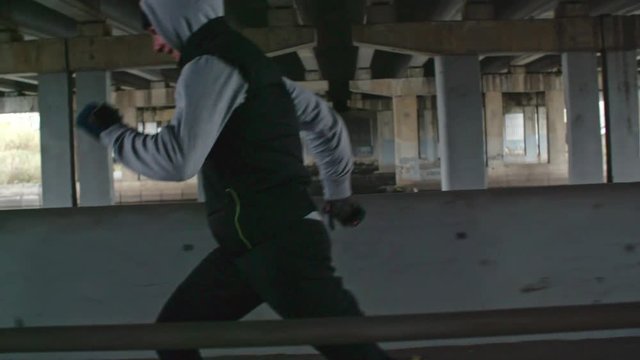 Tracking with slow motion of young sporty man running along pavement under bridge on cold day